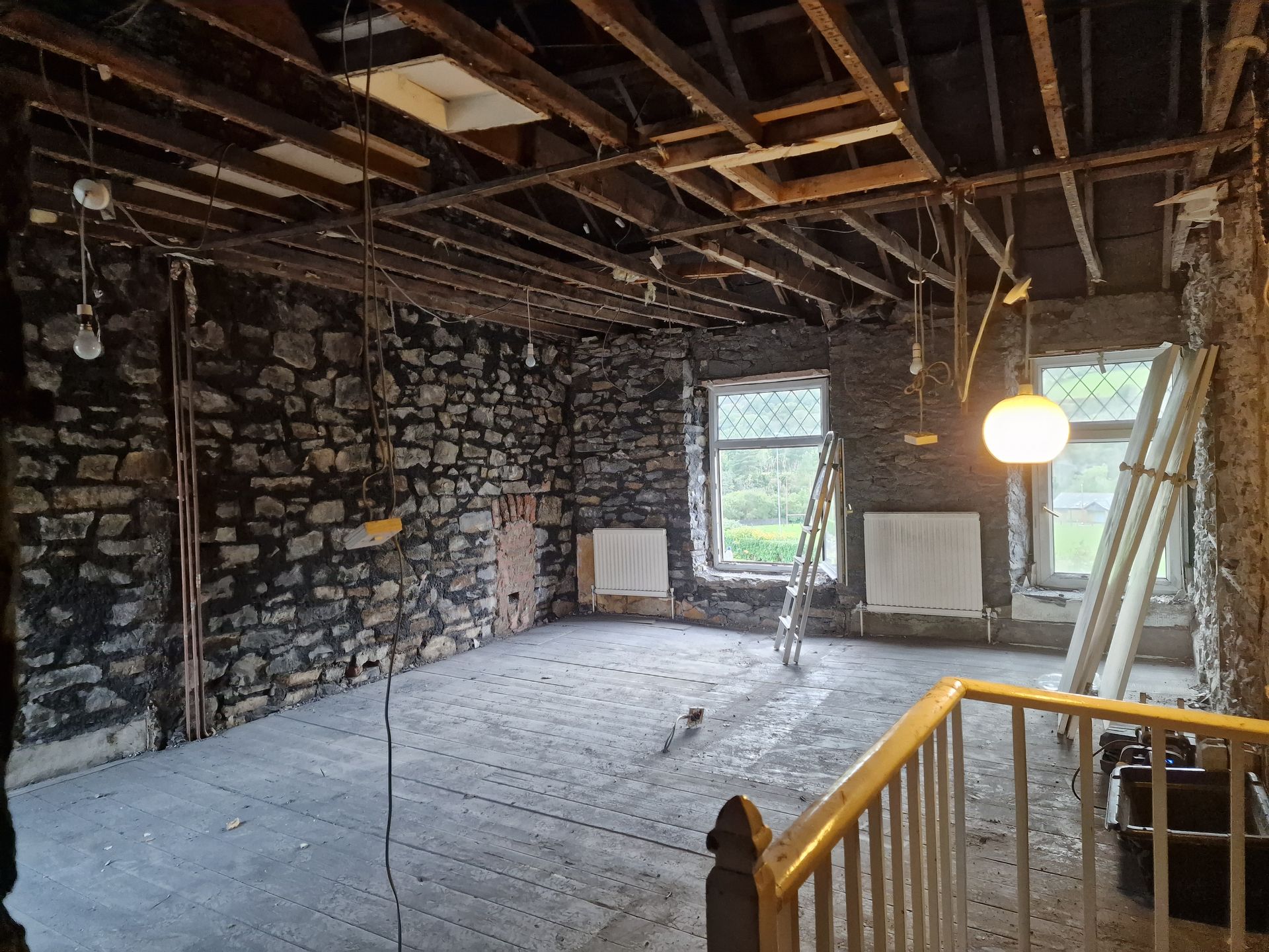 Renovation in progress: exposed stone walls and ceiling beams, two windows, and a partially constructed staircase inside a room.