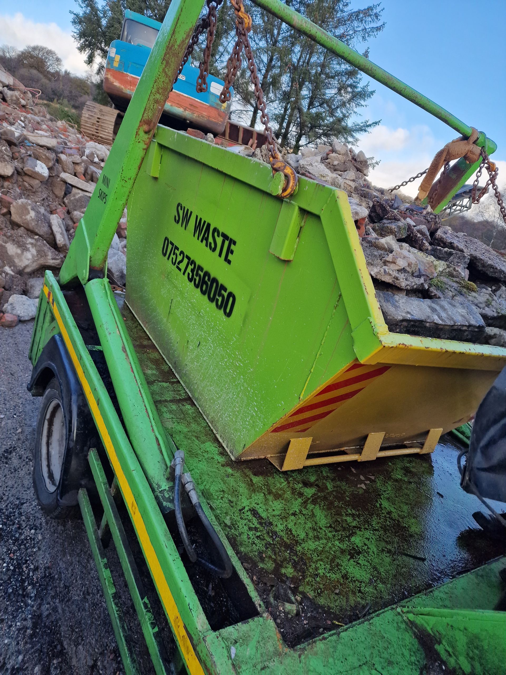 A green skip filled with debris being lifted by a truck. The skip is labeled