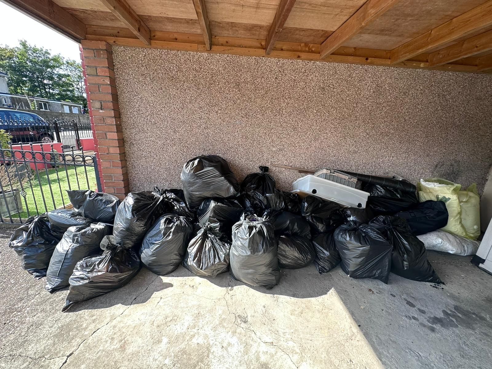 Pile of black trash bags and other discarded items under a covered area, against a textured wall and brick pillar.