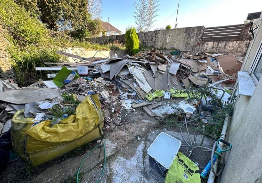 A cluttered backyard with construction debris, including yellow bags, and a white container. Green plants and a concrete wall are in the background.