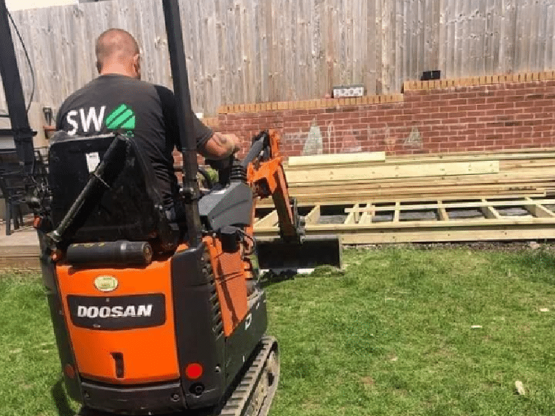 A man operates an orange Doosan excavator on a grassy lawn. Construction materials are nearby.