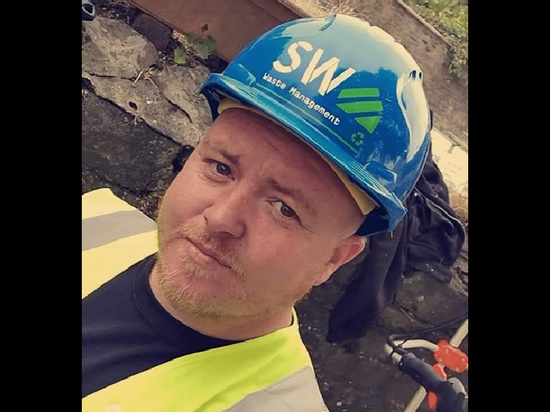 Man with red hair wearing a blue hard hat and safety vest, outdoors. He is looking at the camera with a neutral expression.