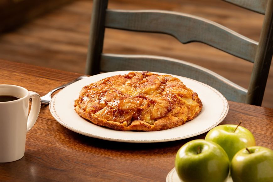 Apple pie on a plate, cup of coffee, and green apples on a wooden table, with a chair in the background.