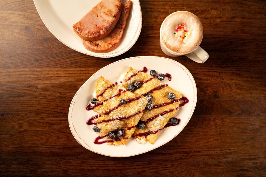 Crepes with blueberries and syrup on a plate, ham, and a cappuccino on a wooden table.