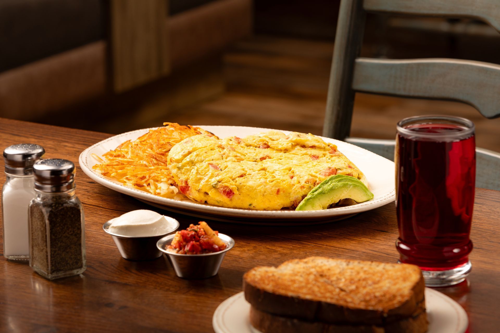 Breakfast plate with omelet, hash browns, toast, juice, and condiments on a wooden table.
