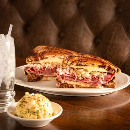 Reuben sandwich on a plate with potato salad and a drink, brown background.