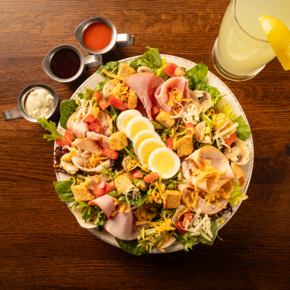 Salad with ham, eggs, and cheese, served with sauces and lemonade on a wooden table.