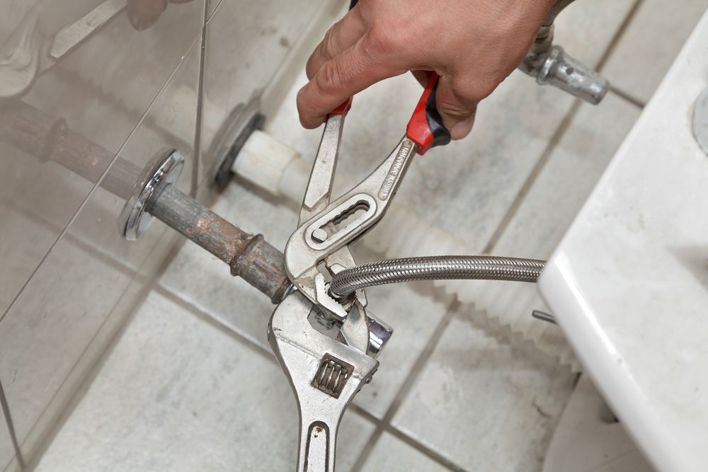 A Person Uses Pliers and a Wrench to Repair Plumbing Under a White Sink on a Tiled Floor — No. 2 Plumbing In Kenmore, QLD