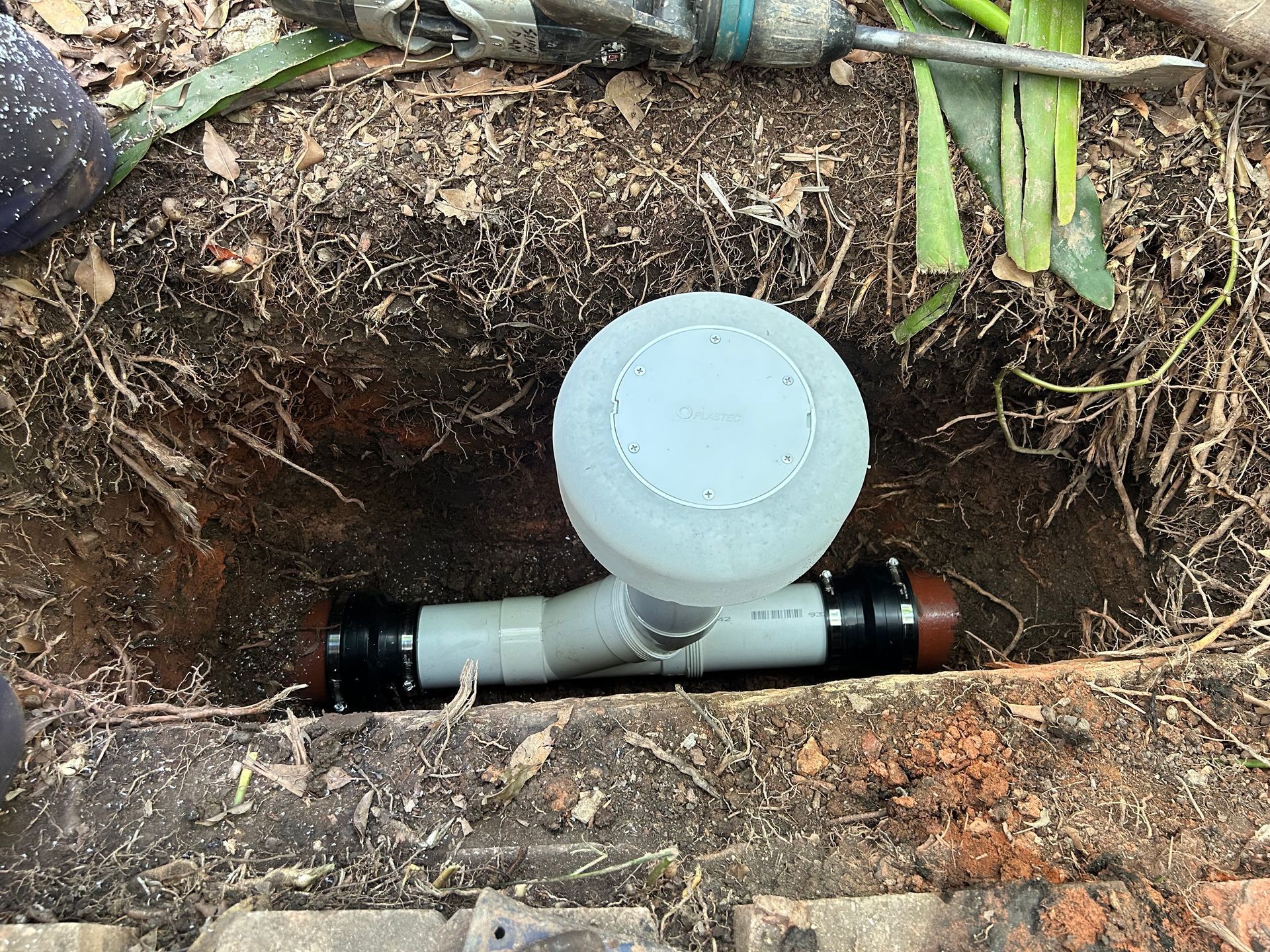 A grey plastic sewer cleanout assembly with a round access cap, installed between pipes in a trench dug into dirt — No. 2 Plumbing In Kenmore, QLD