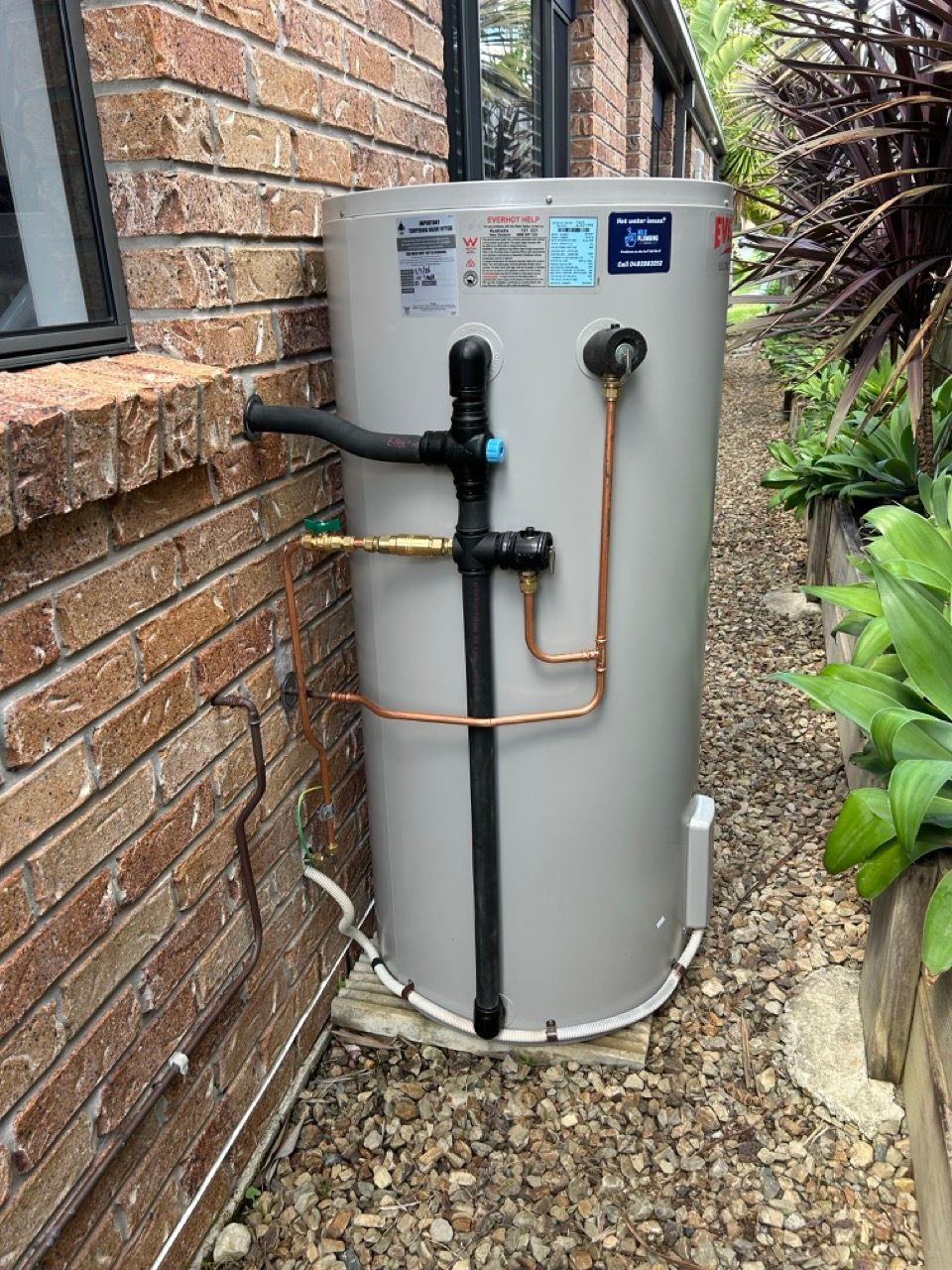 A grey cylindrical outdoor water heater unit positioned against a brick wall on gravel ground — No. 2 Plumbing In Kenmore, QLD