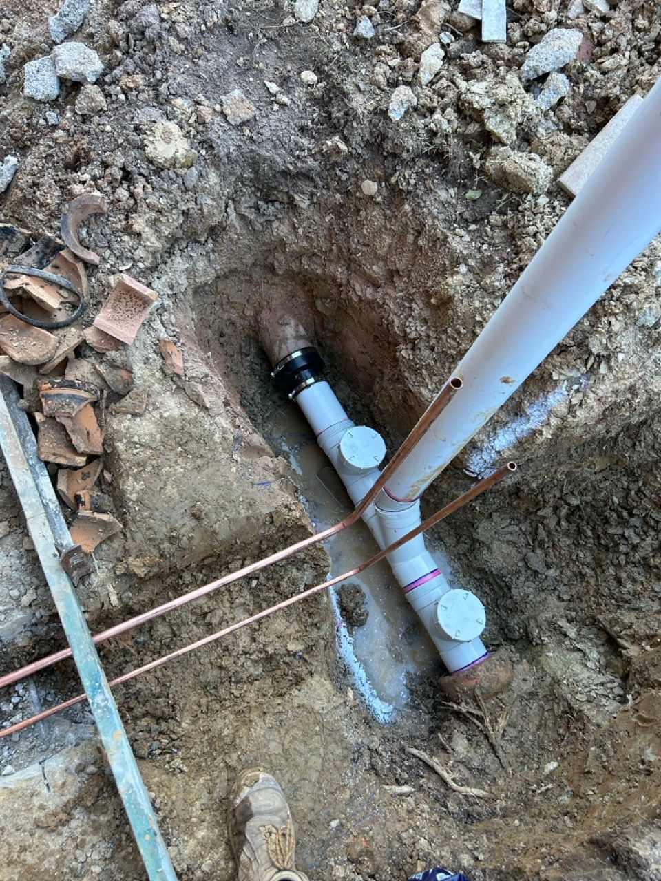 A white PVC drainage pipe with two cleanouts sits in a dirt trench, connected to a dark pipe via a rubber coupling — No. 2 Plumbing In Kenmore, QLD