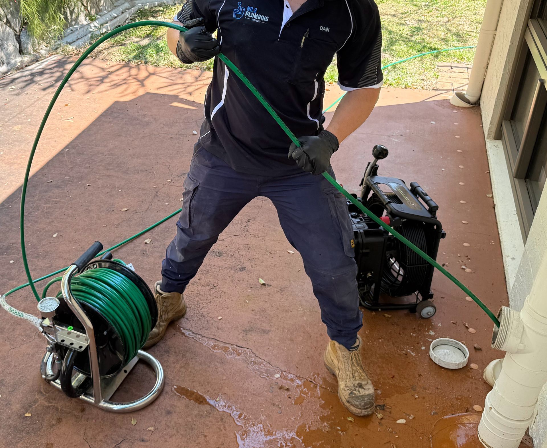 Plumber uses a drain cleaning machine near a building. He wears black gloves, and a green hose is visible. — No. 2 Plumbing In Kenmore, QLD