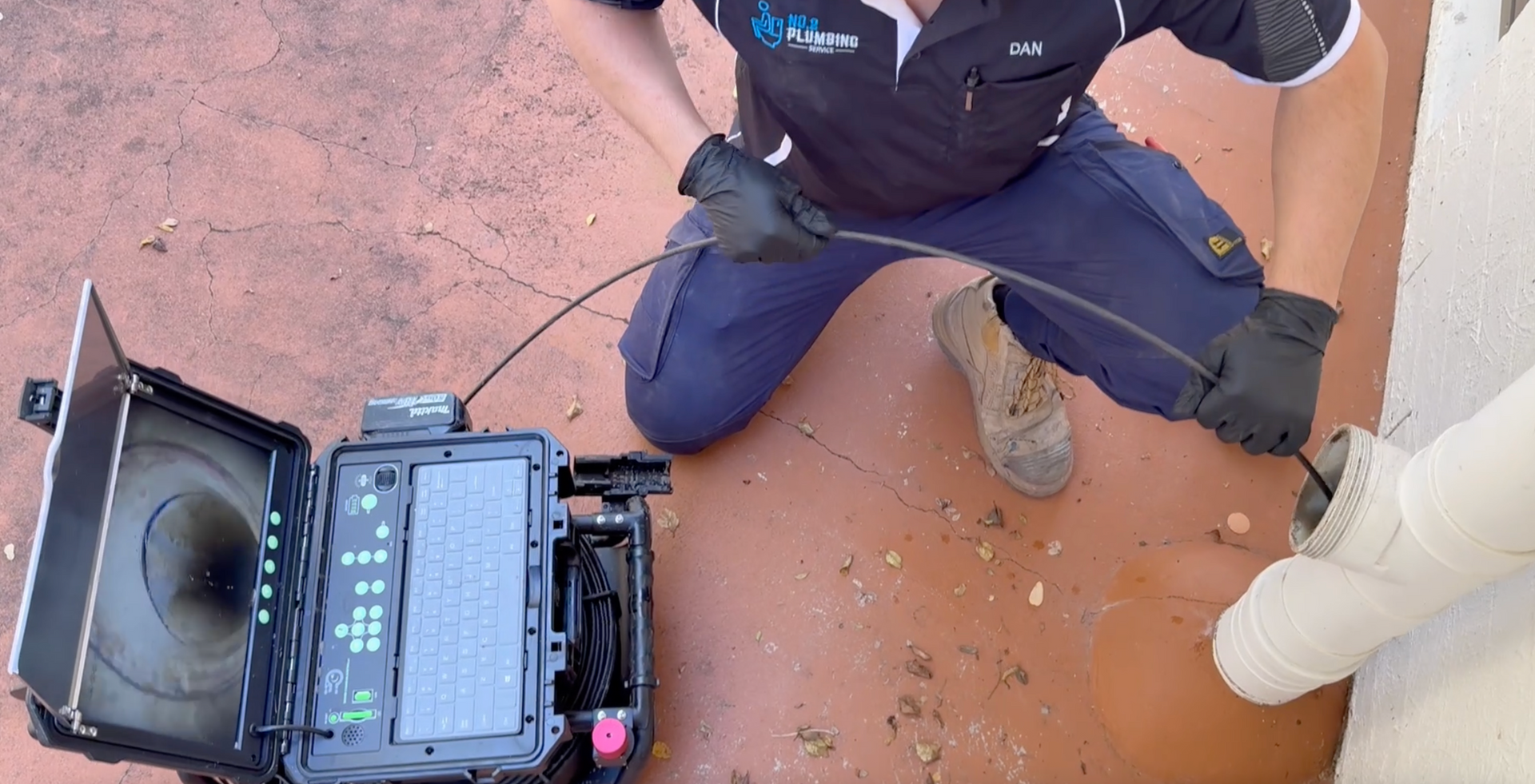 Plumber using camera equipment to inspect a pipe. He kneels on a red concrete surface, wearing gloves and dark work clothes. — No. 2 Plumbing In Kenmore, QLD