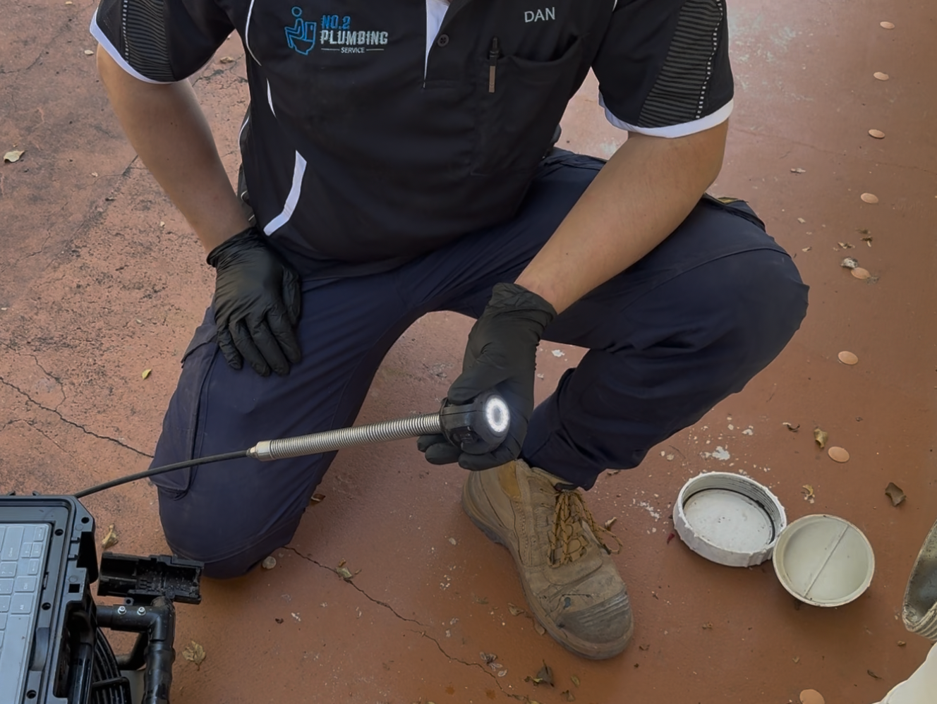Person in work attire inspecting a surface with a tool, outdoors, with containers of substance visible. — No. 2 Plumbing In Kenmore, QLD