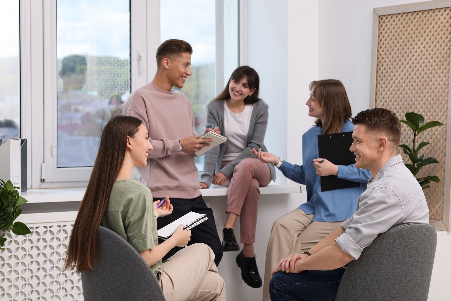 Group of people in casual wear talking and smiling in a well-lit room; some are holding notebooks.
