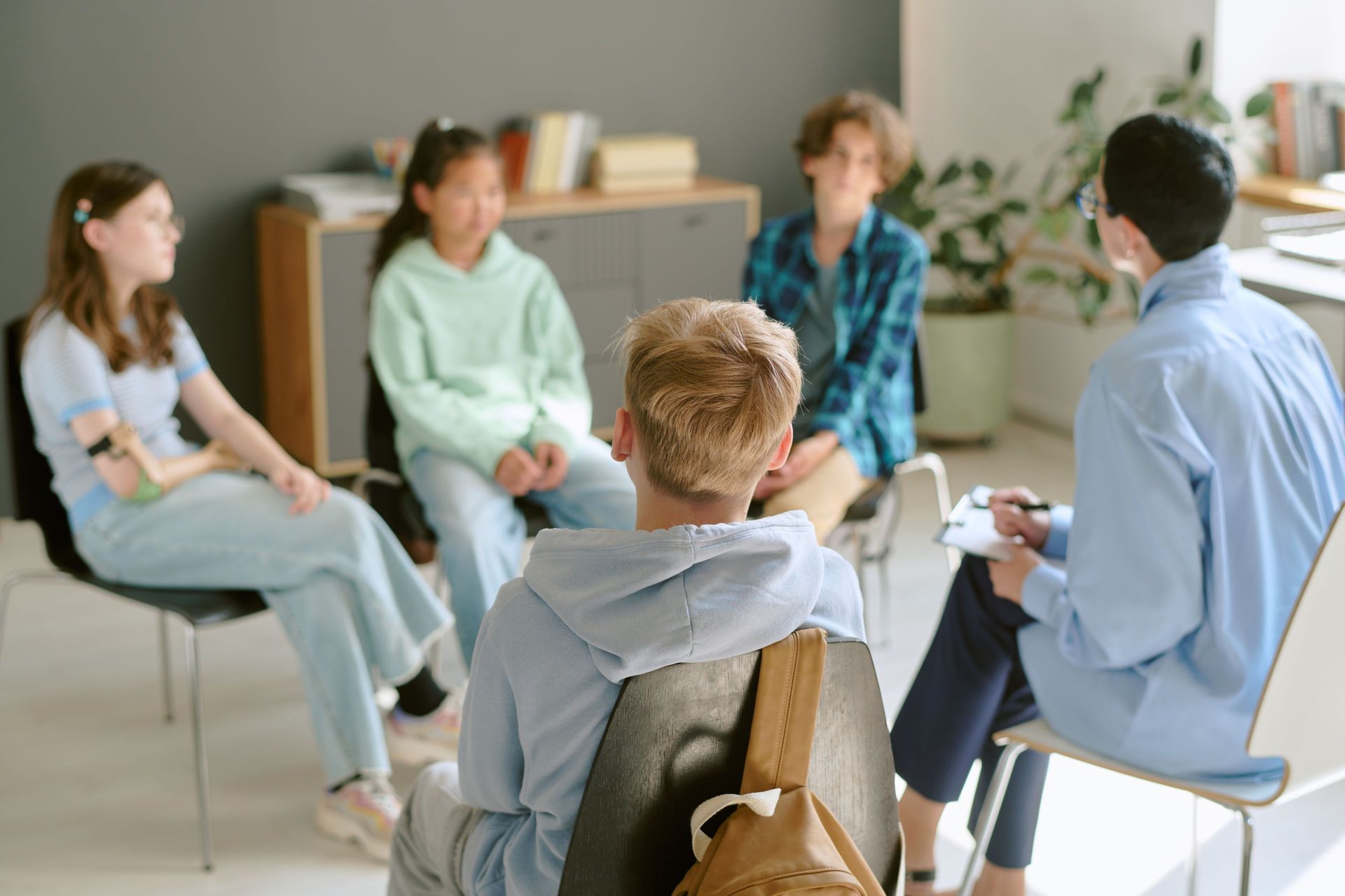 Group of teens in a circle with a person holding a clipboard, discussing in a room.