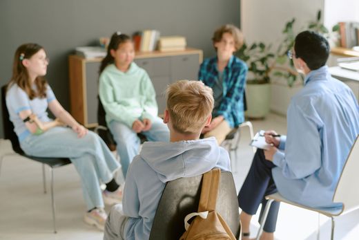 A group of teens in a circle with a counselor, discussing in an office setting.