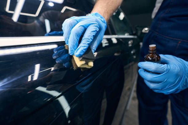 A person applying ceramic coating to a black car panel with a sponge and a small bottle. They are wearing blue gloves.