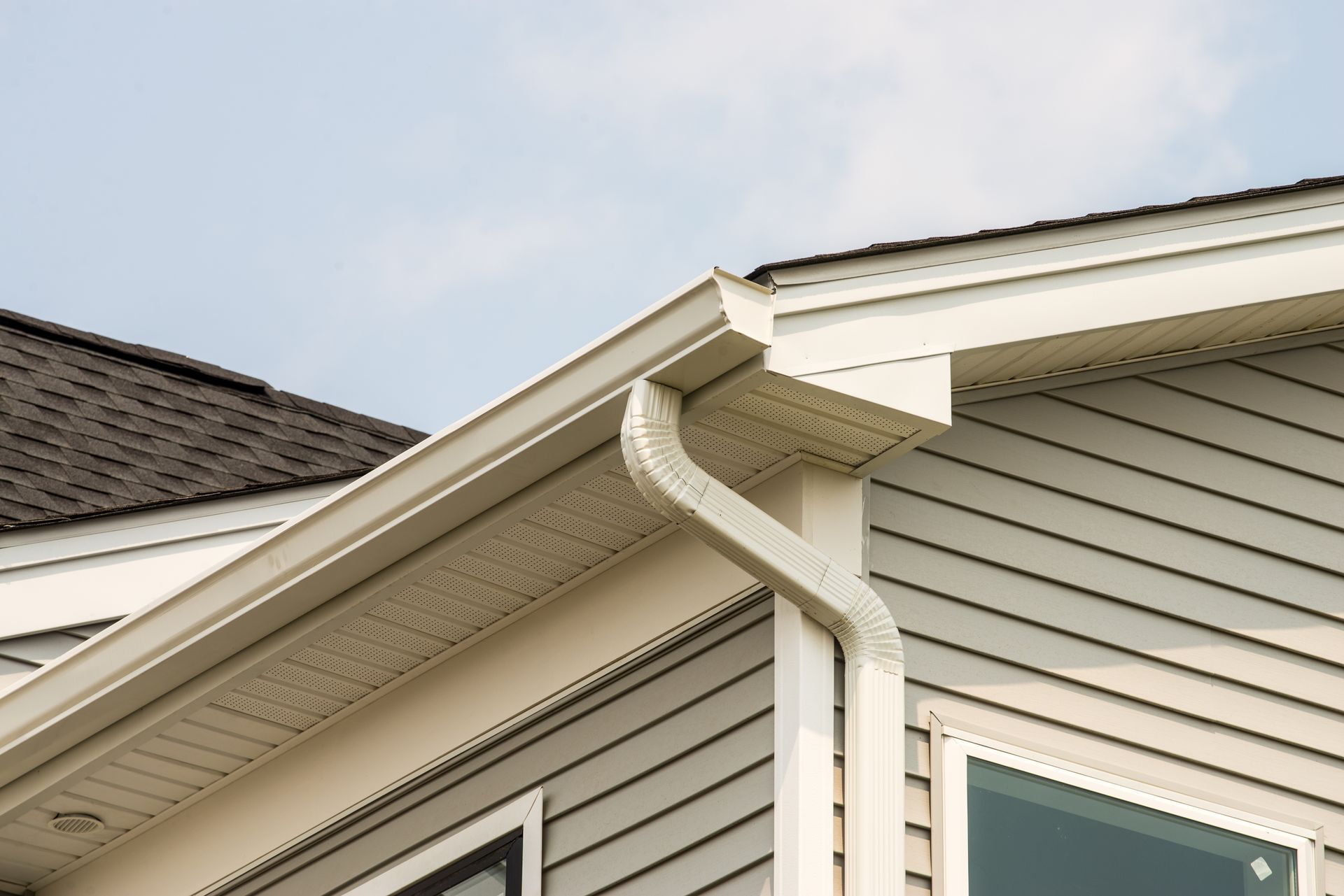 Beige rain gutter on a house with gray siding and a dark roof under a blue sky.