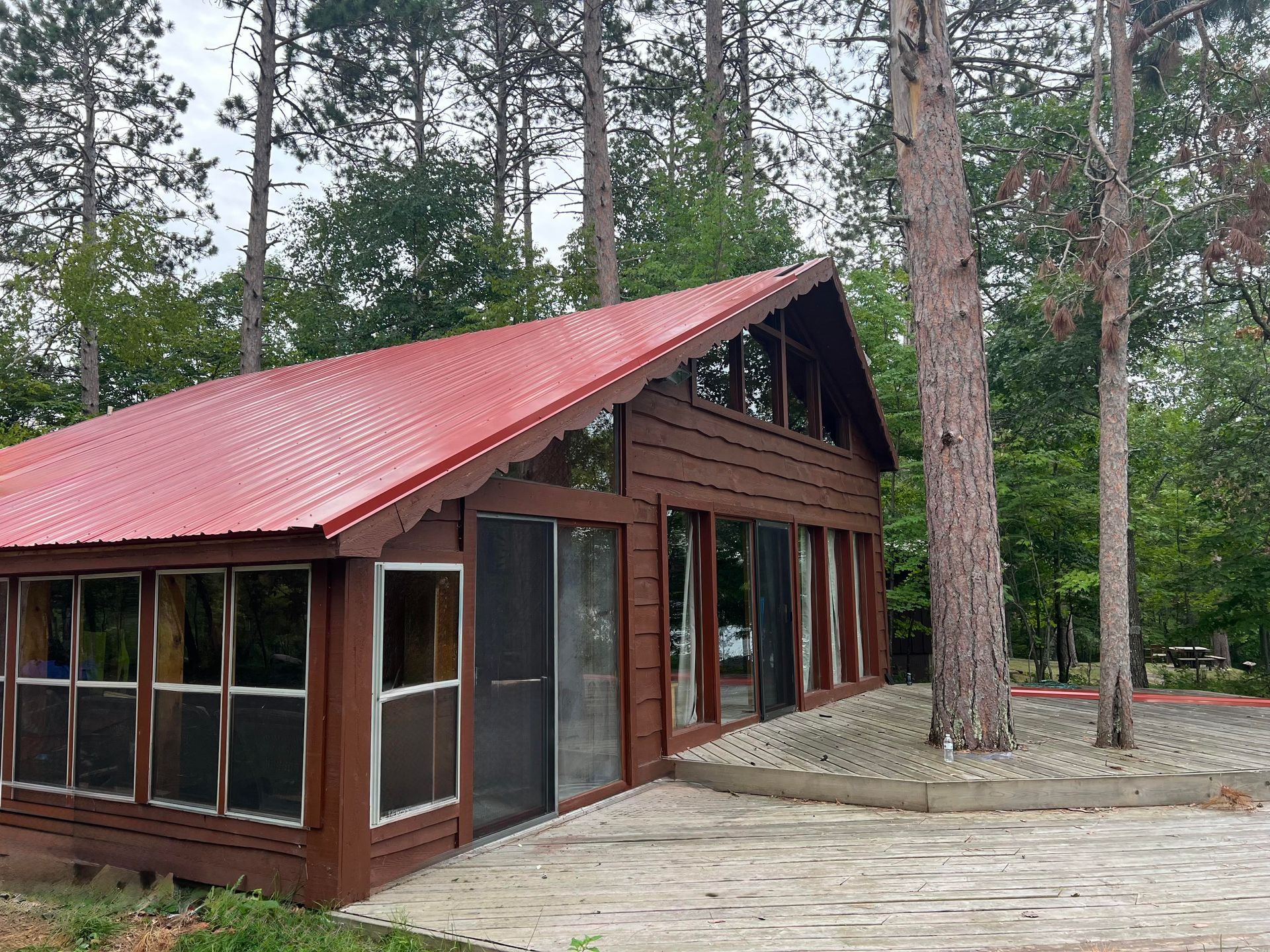 Brown cabin with a red roof and large windows, surrounded by trees. A concrete patio is in front.