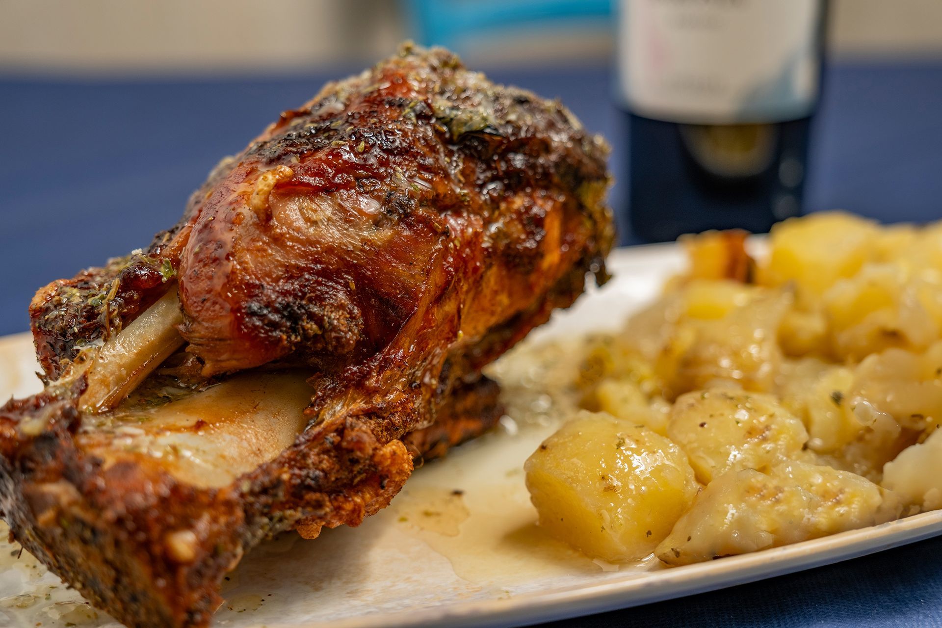 Spalla di maiale arrosto con patate, su un piatto bianco, tovaglia blu, bottiglia di vino sullo sfondo.