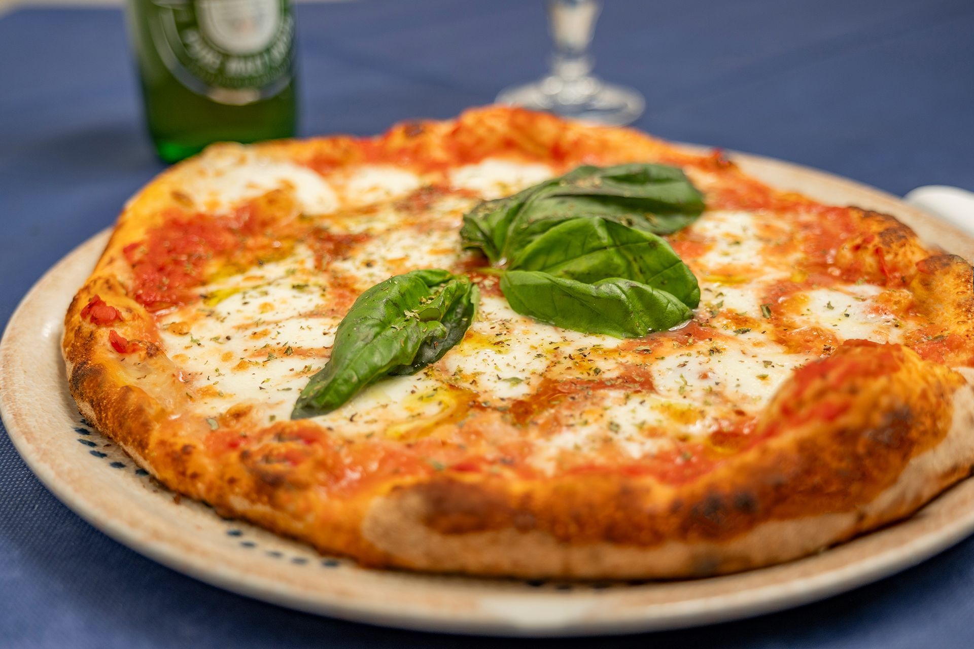 Pizza su un piatto, condita con formaggio e basilico. Sullo sfondo una bottiglia di birra.
