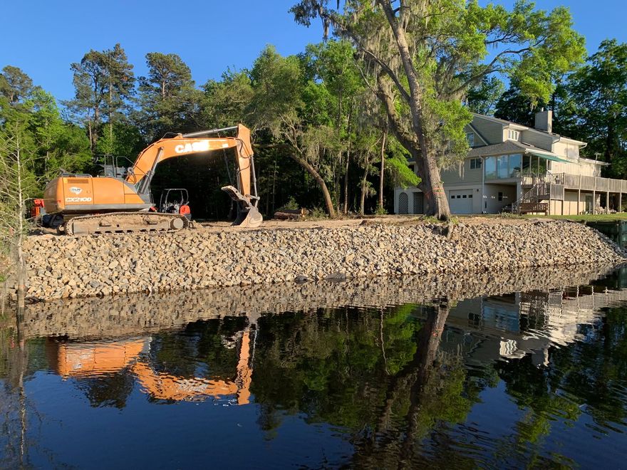 A large excavator is sitting on the shore of a lake next to a house.