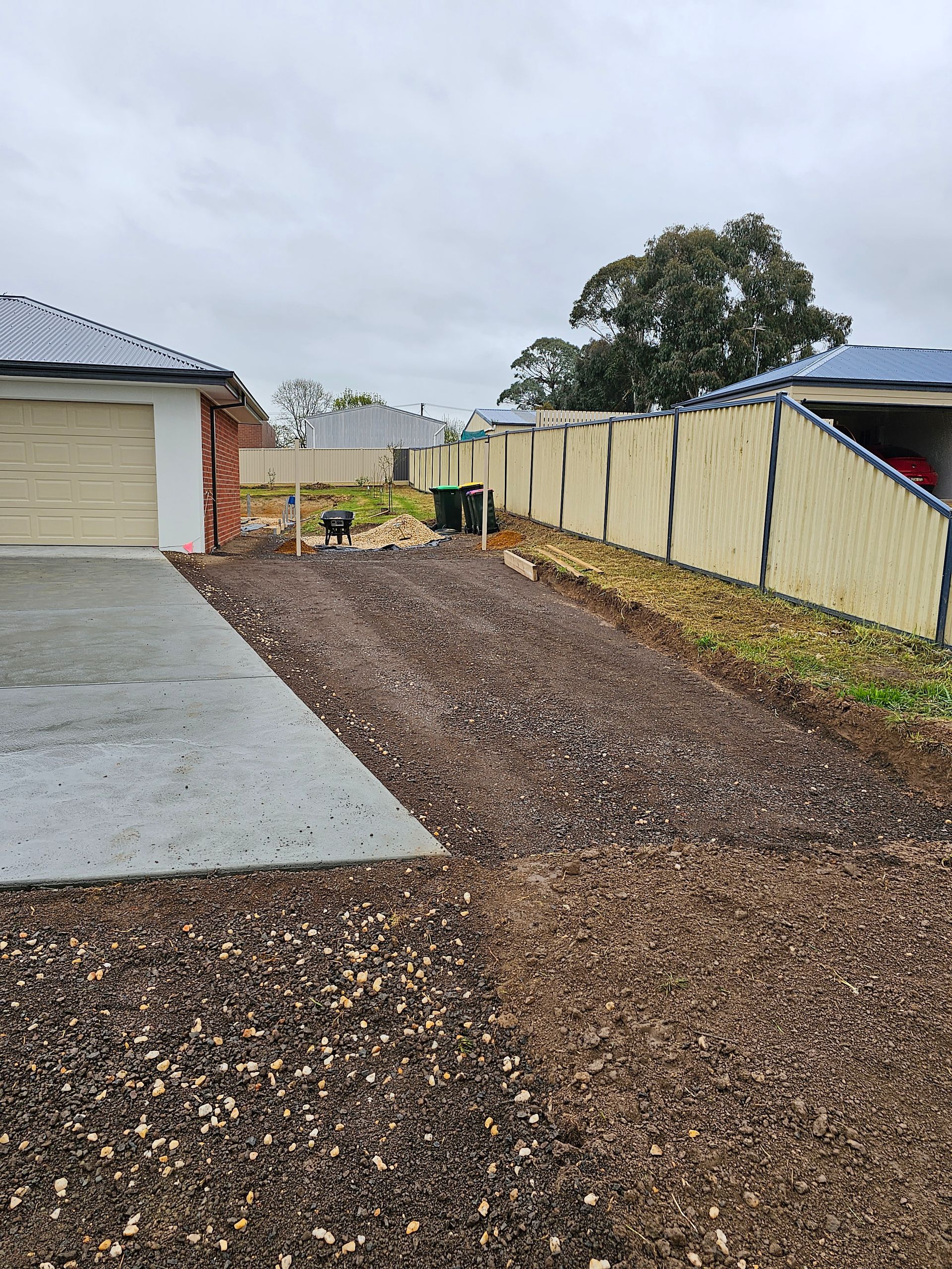 A gravel-covered yard next to a concrete driveway, with a fence and houses in the background under a cloudy sky.