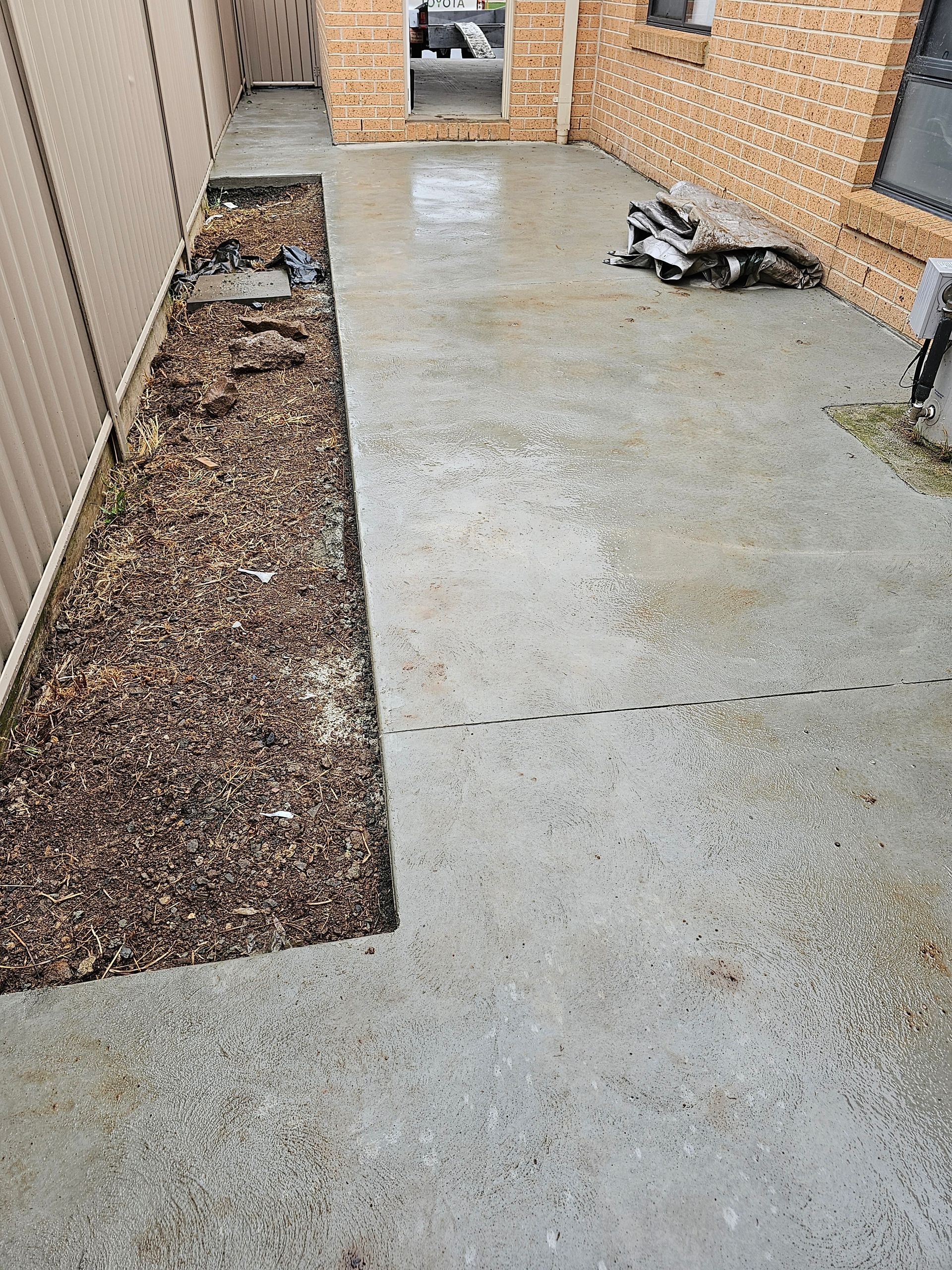 Concrete patio next to a narrow garden bed filled with mulch, bordered by a beige fence and brick wall.