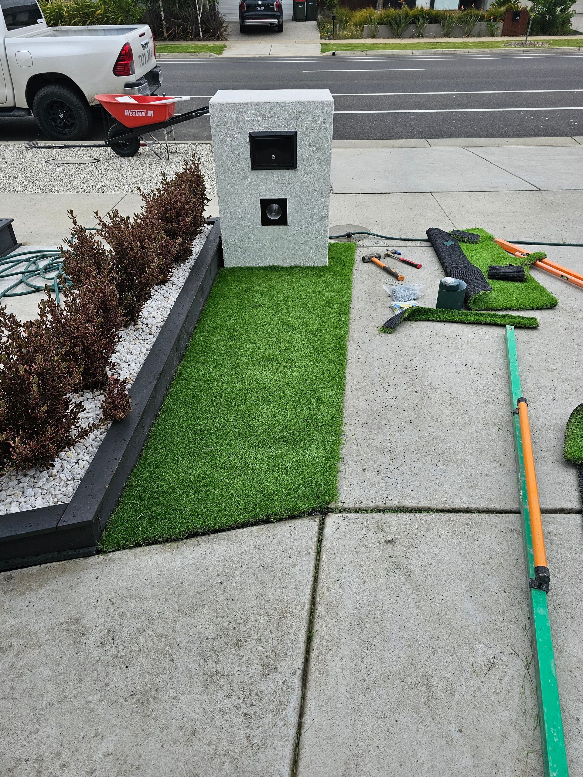 A person installs artificial turf near a mailbox and a planter box. The project is on a concrete driveway next to a street.
