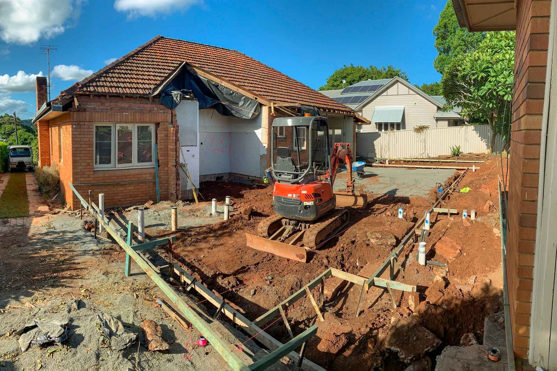 Construction site with a small excavator in a dirt trench next to a brick house under renovation. Sunny day, clear sky.
