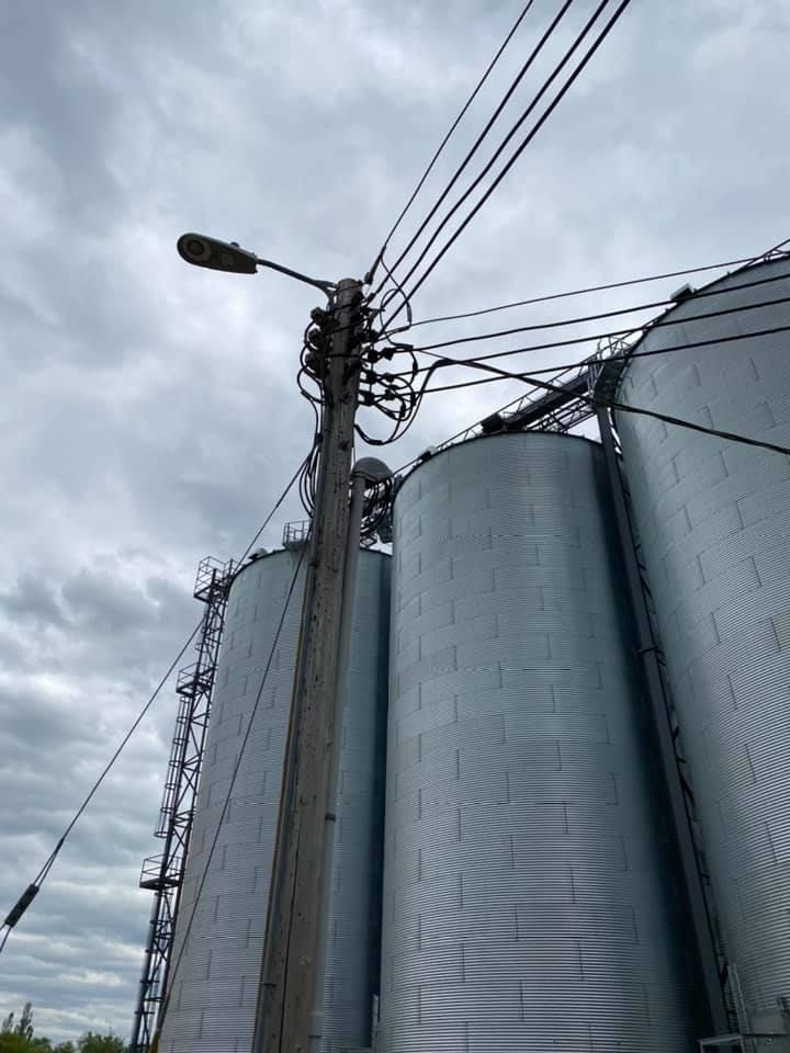 Power lines and a street lamp atop a wooden pole beside tall metal silos under a cloudy sky.