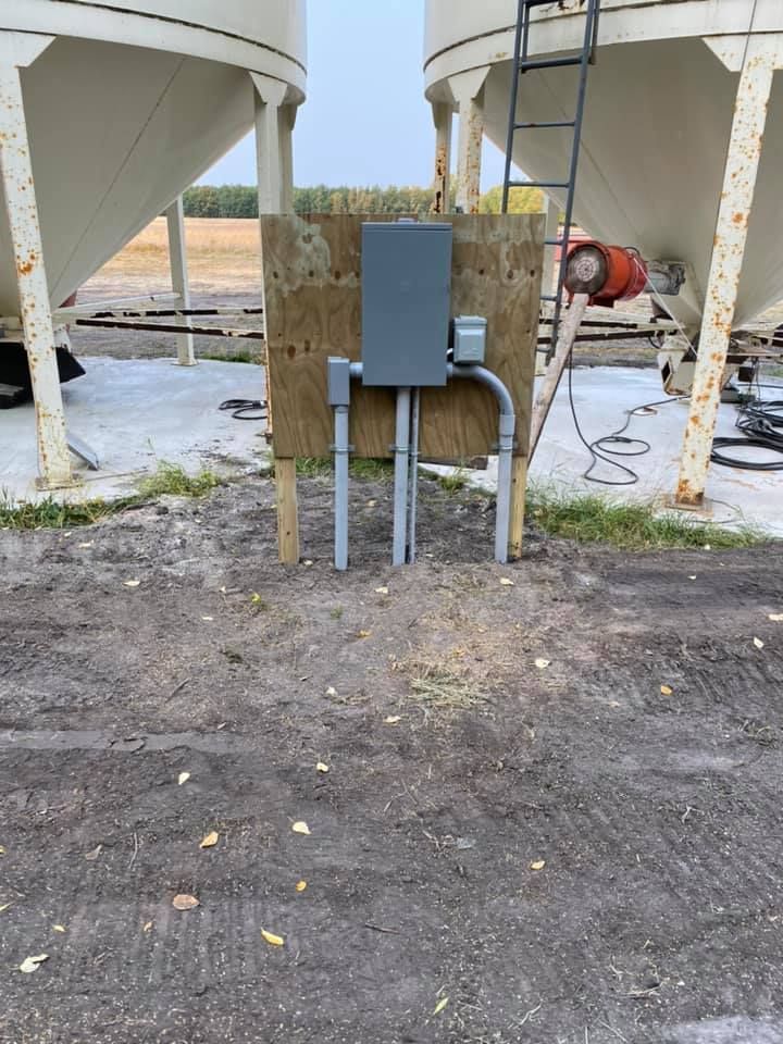 Electrical box on a plywood board between two large grain silos, gray pipes, outdoor setting.