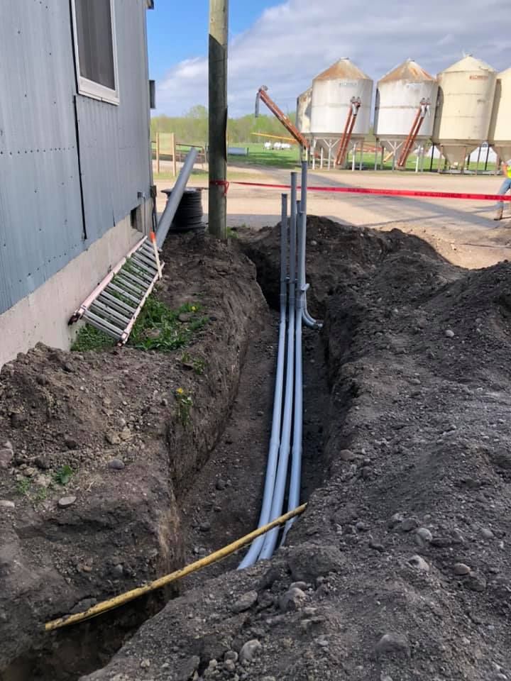 Trench with electrical conduit running alongside a building and utility pole, in front of grain silos.