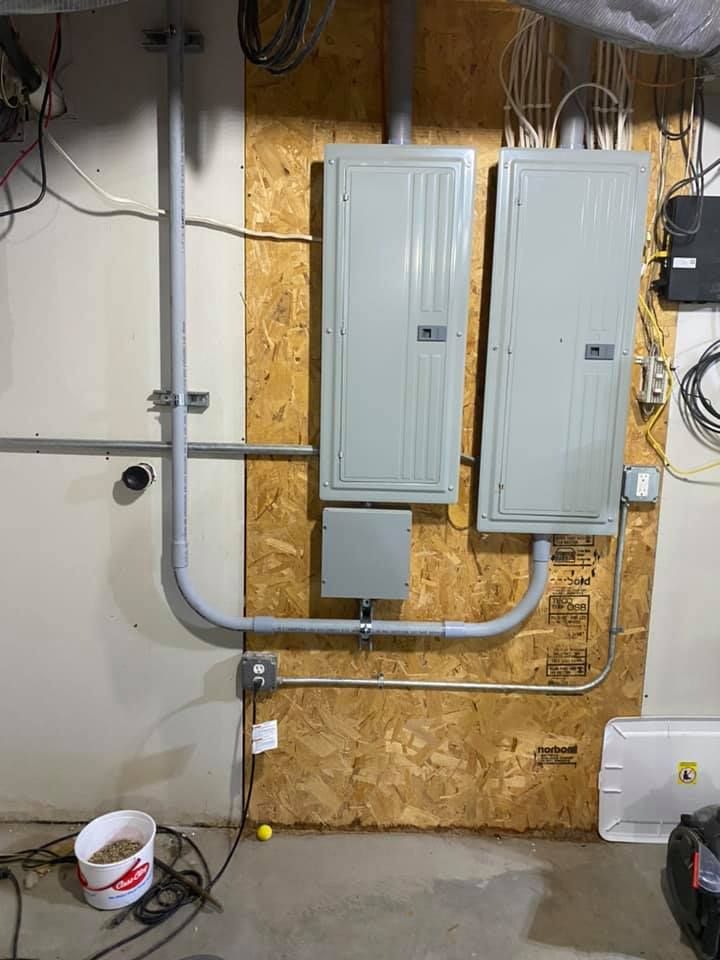 Electrical panels and conduit mounted on plywood in a basement.
