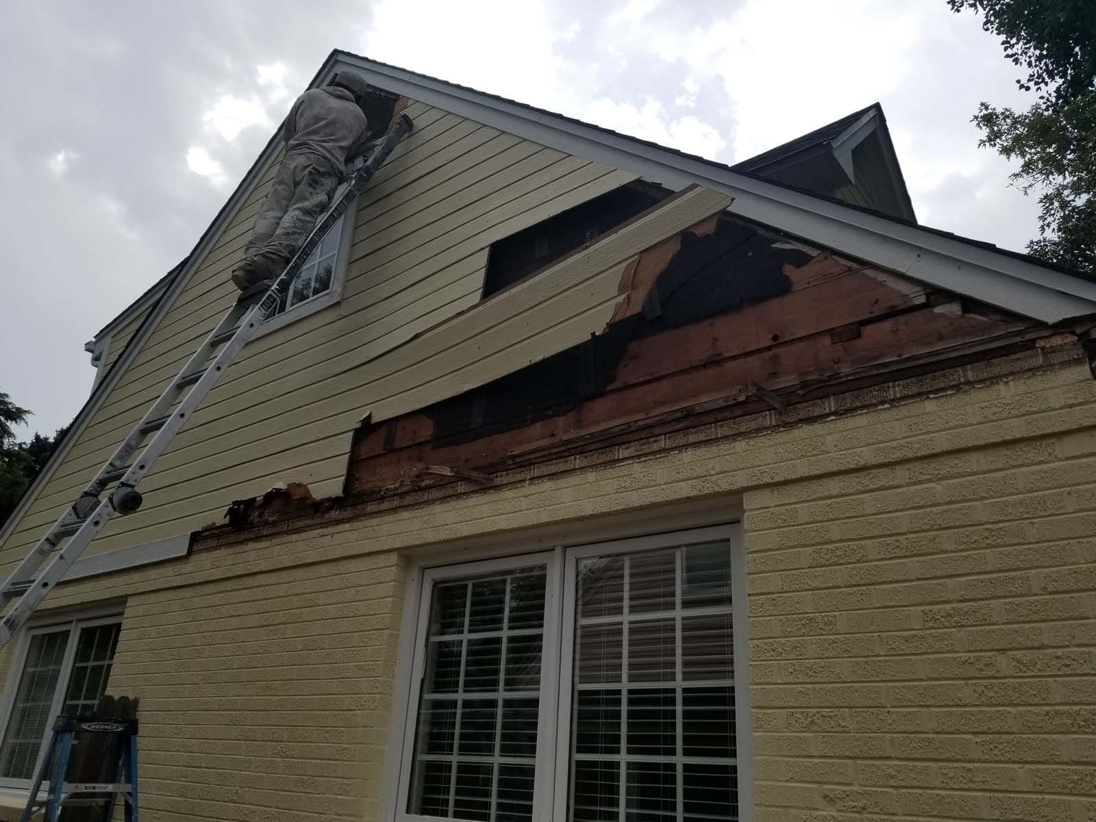 House exterior damage: missing siding, exposing brick, ladder against wall; cloudy sky.