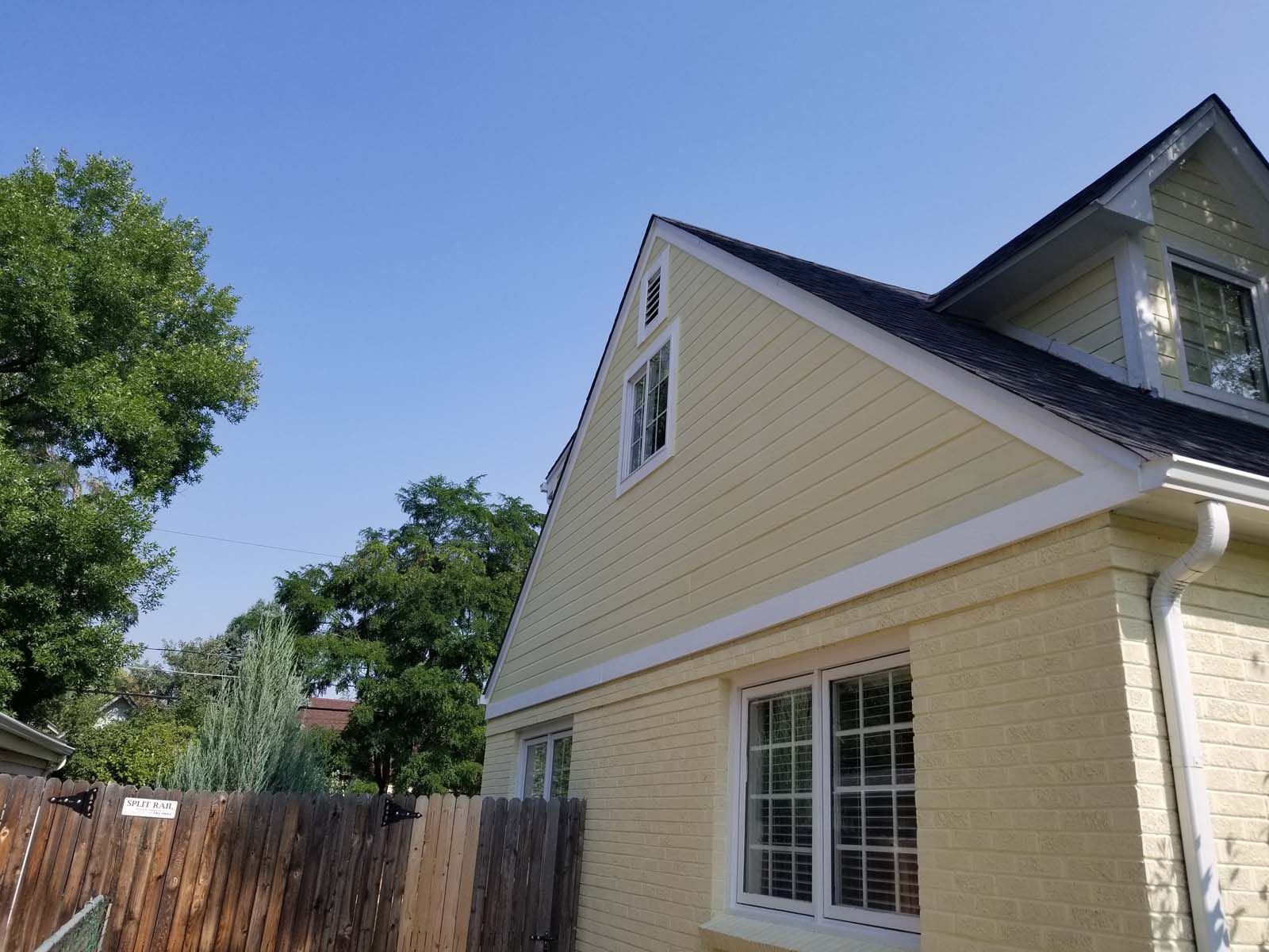 Yellow house with black roof against a blue sky, with a wooden fence and trees in the background.