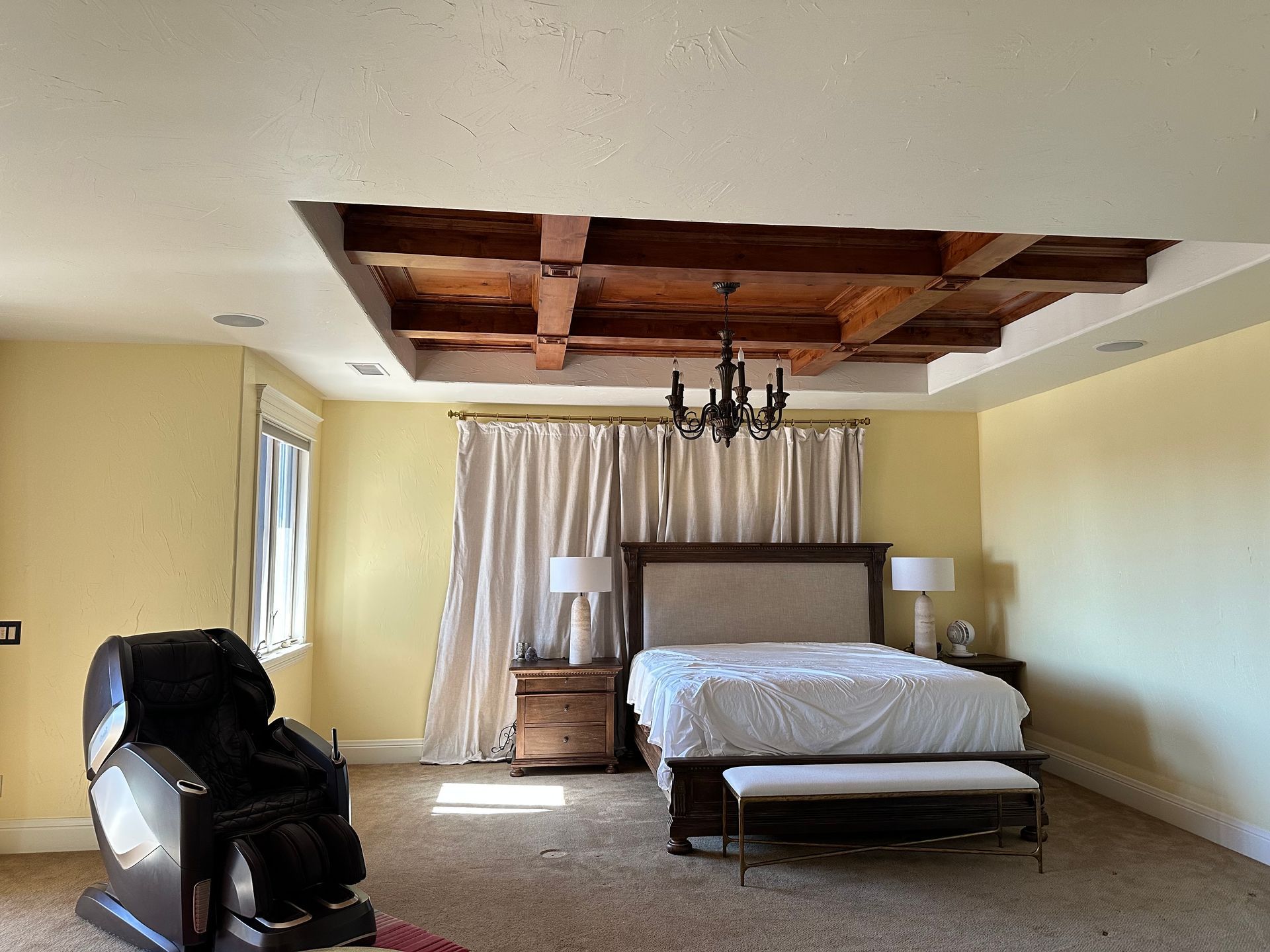 Bedroom with a wood-beamed ceiling, bed, massage chair, and curtains; beige walls.