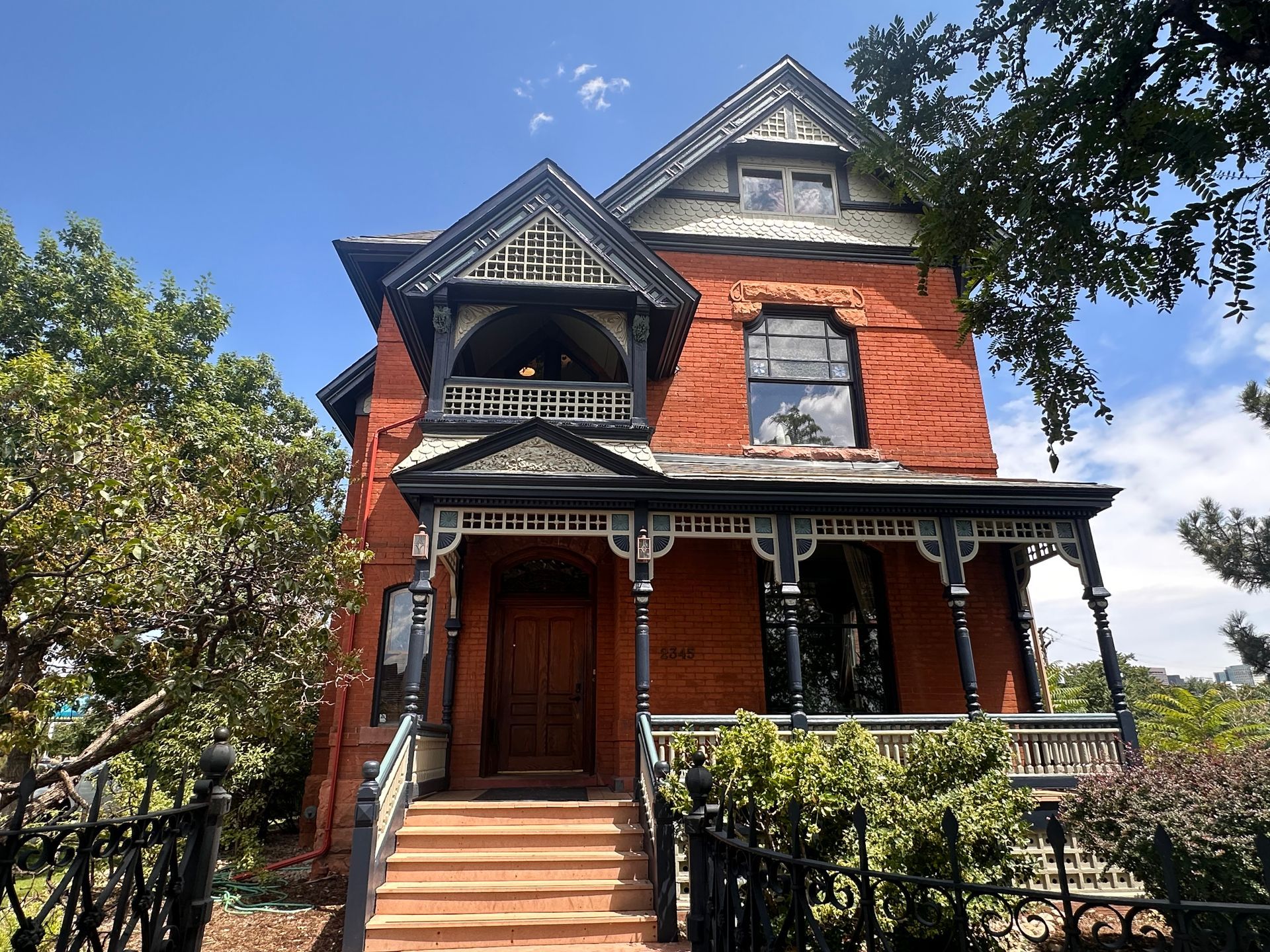 Red brick Victorian house with ornate porch and balcony, steps leading to wooden door, blue sky.