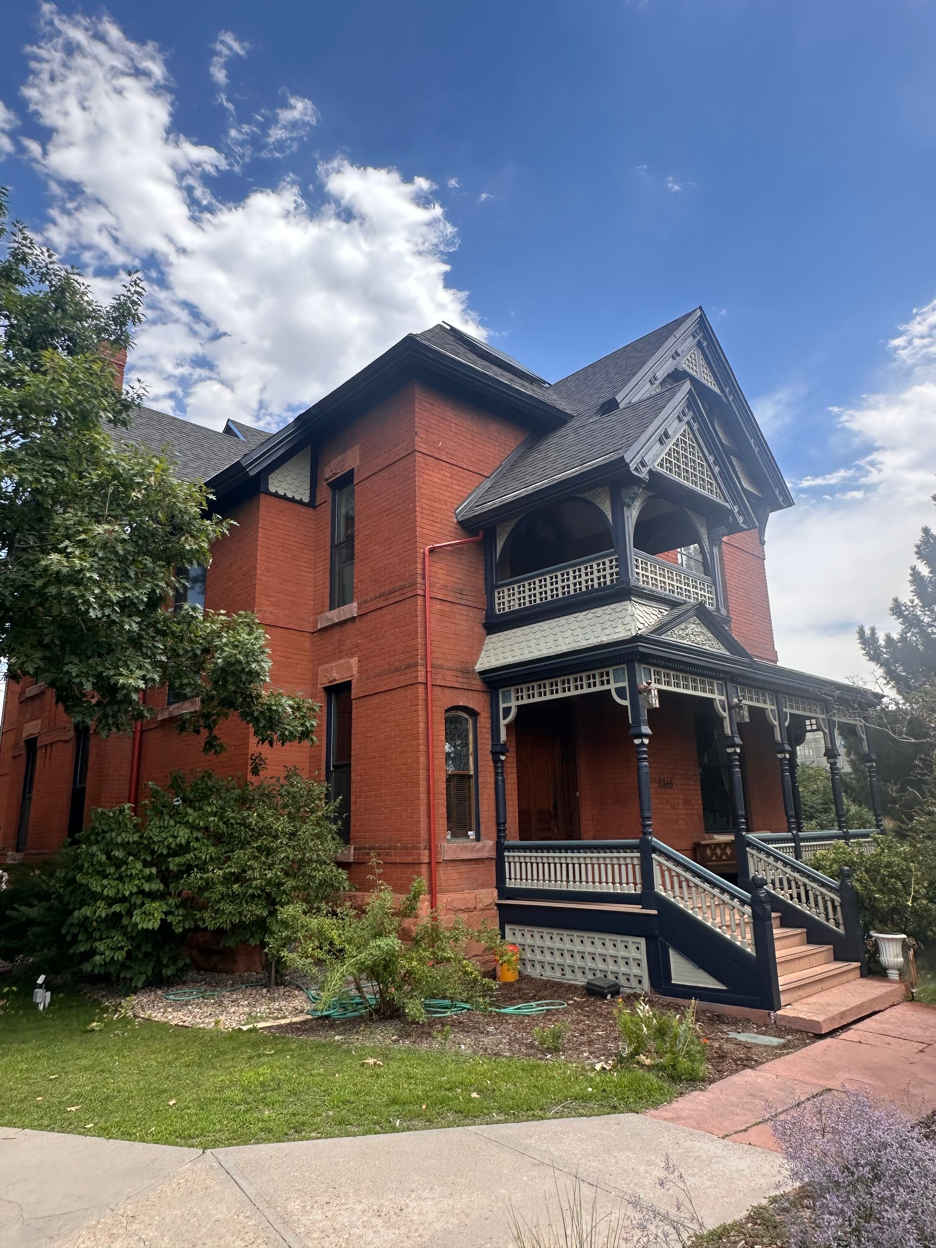 Red brick Victorian house with ornate dark trim, porch, and green landscaping under a blue sky.