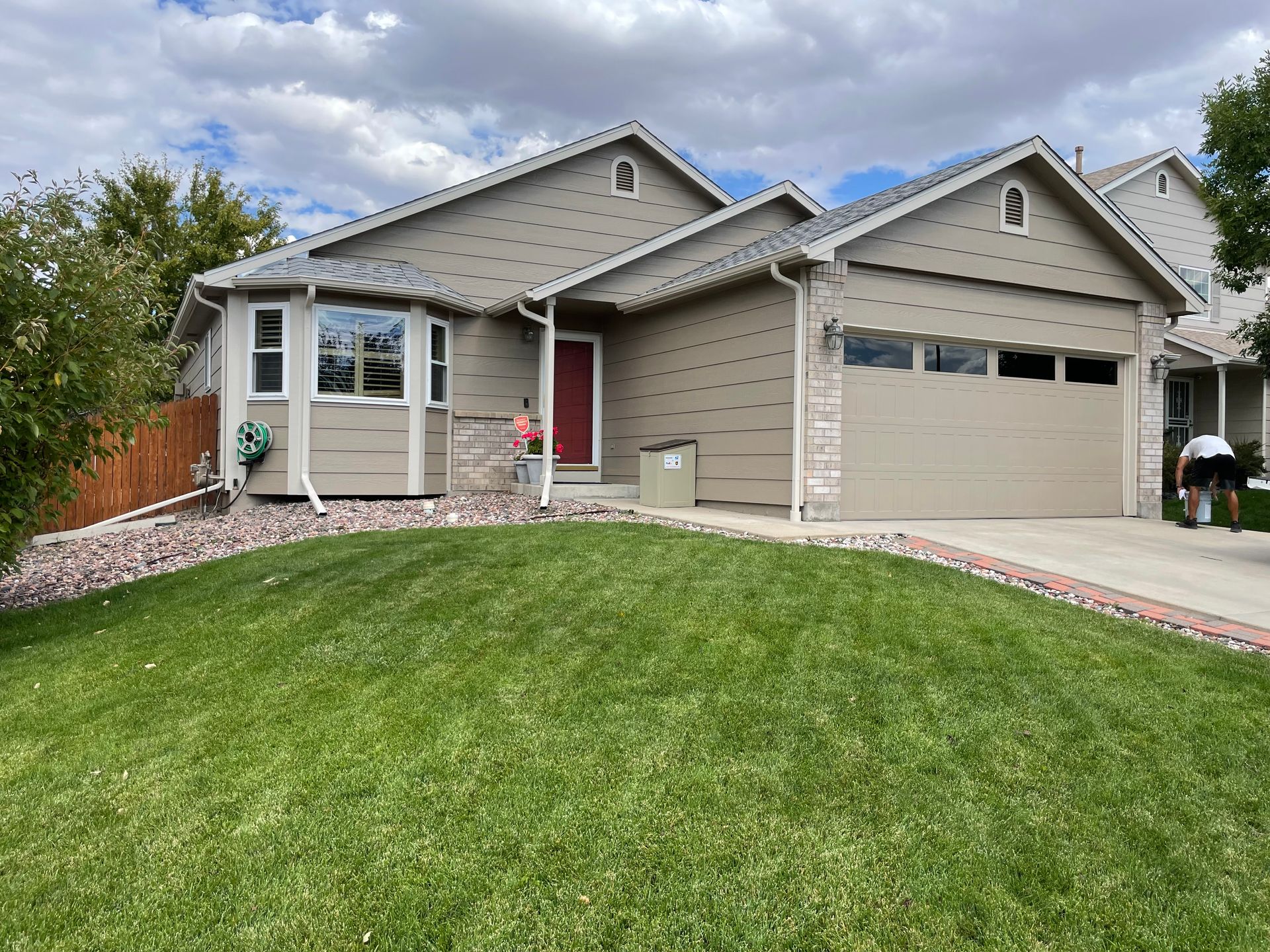 Tan house with a red door, green lawn, and a person washing a car outside the garage.