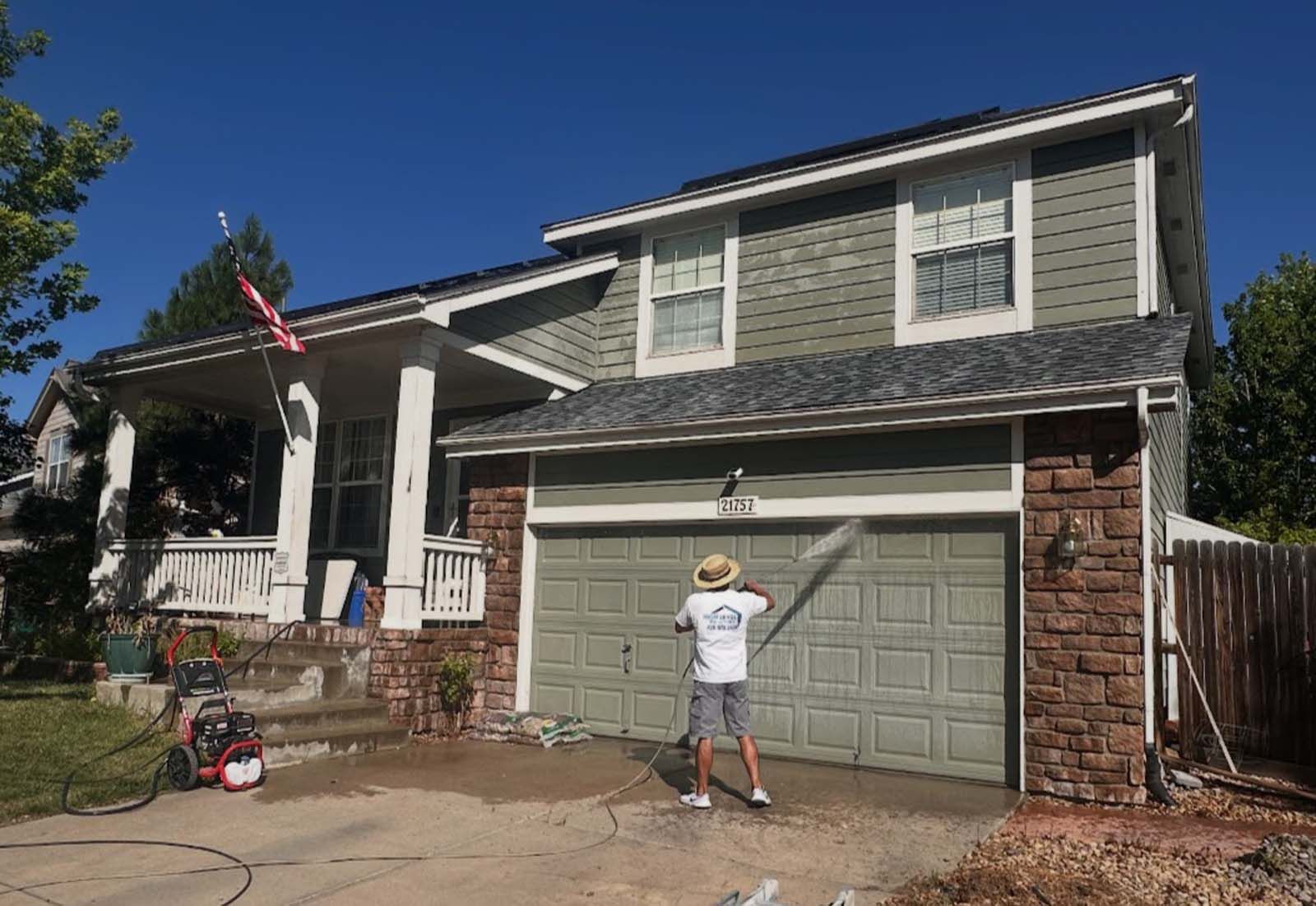 Man power washing a two-story house with green siding, garage door, and a red and white pressure washer.