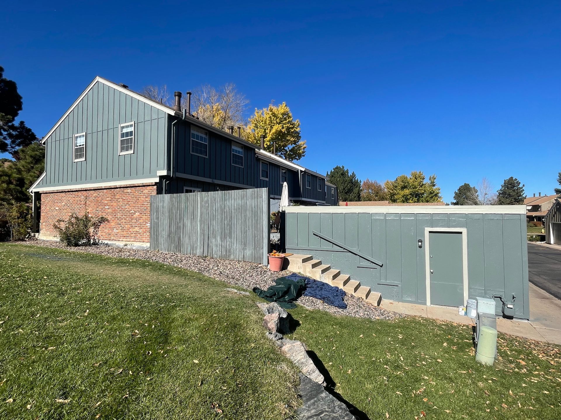 Two-story blue-green building with brick accents, steps, a green door, and a grassy lawn under a clear blue sky.