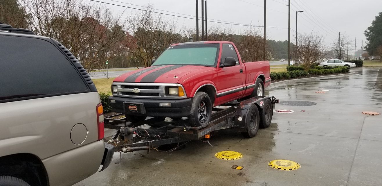 A red truck is being towed by a trailer in a parking lot.