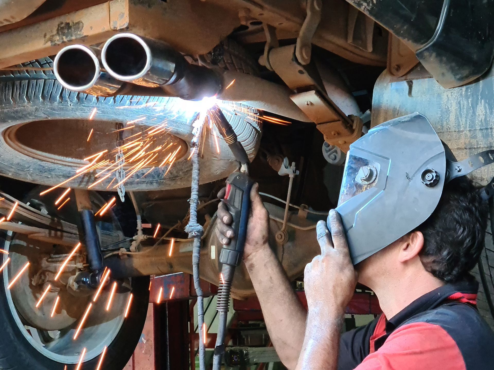A man wearing a welding mask is welding under a car