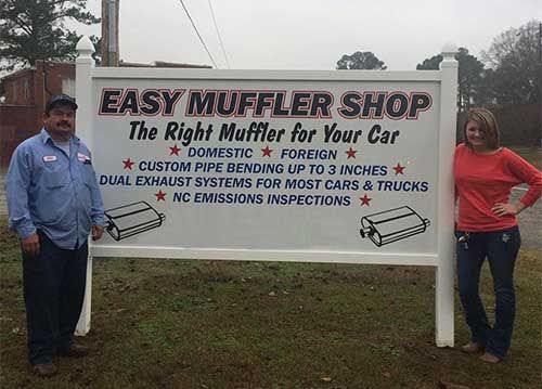 A man and a woman are standing in front of a sign for easy muffler shop.