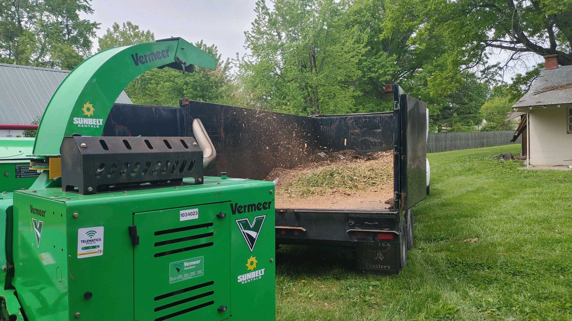 A green chipper is sitting next to a dumpster in a yard.