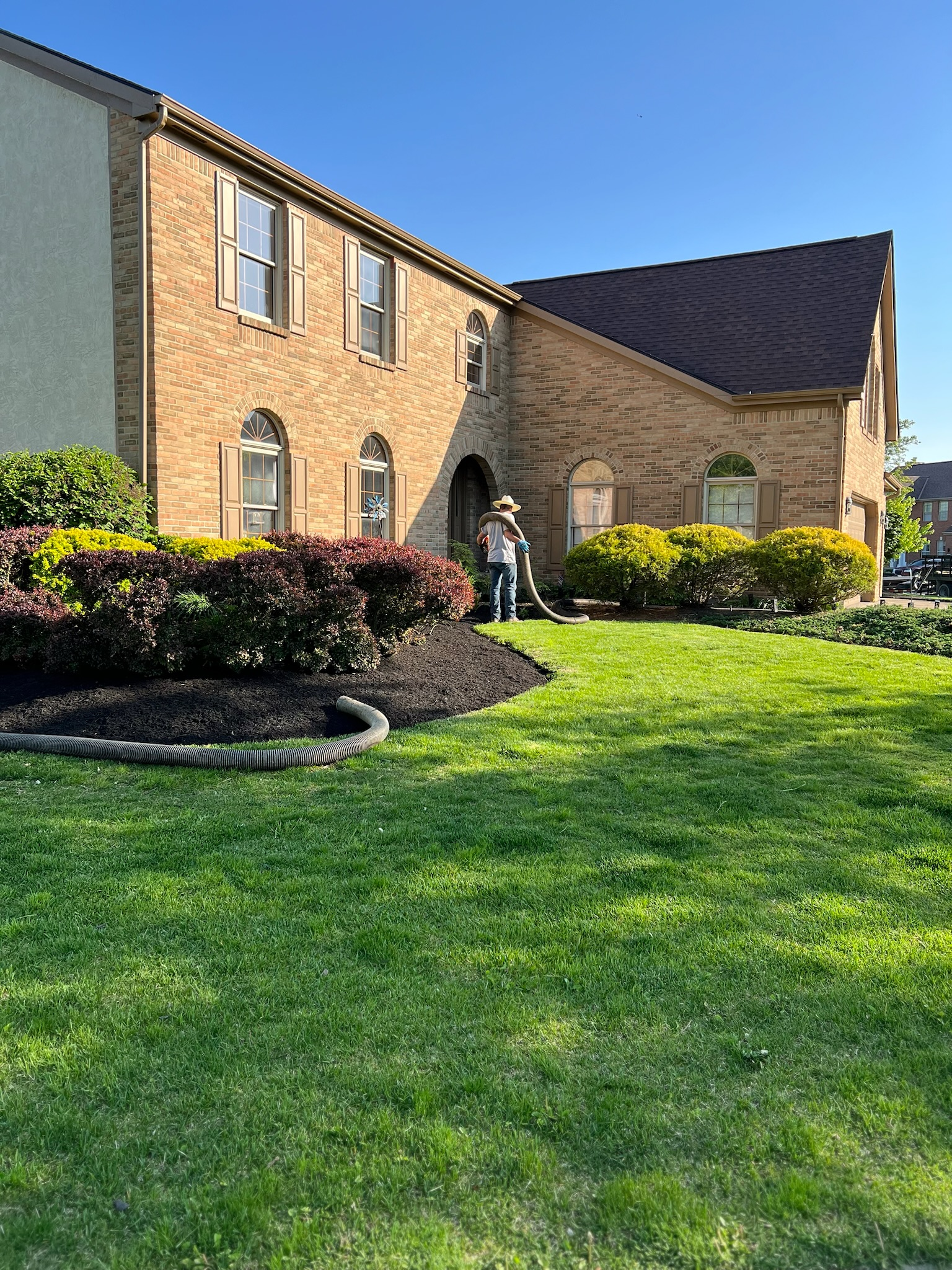 A man is standing in front of a large brick house with a lush green lawn.