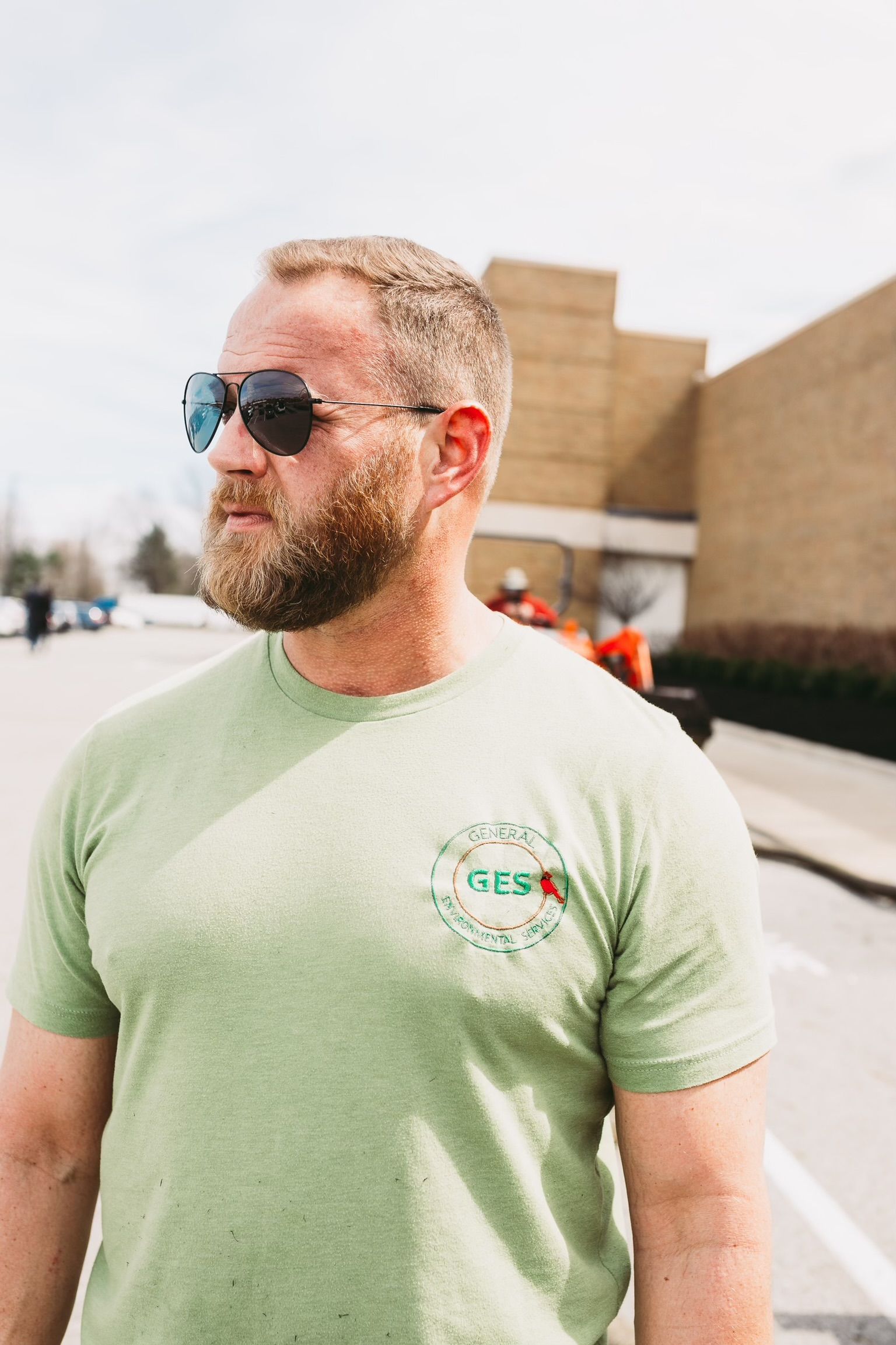 A man with a beard wearing sunglasses and a green shirt is standing in a parking lot.