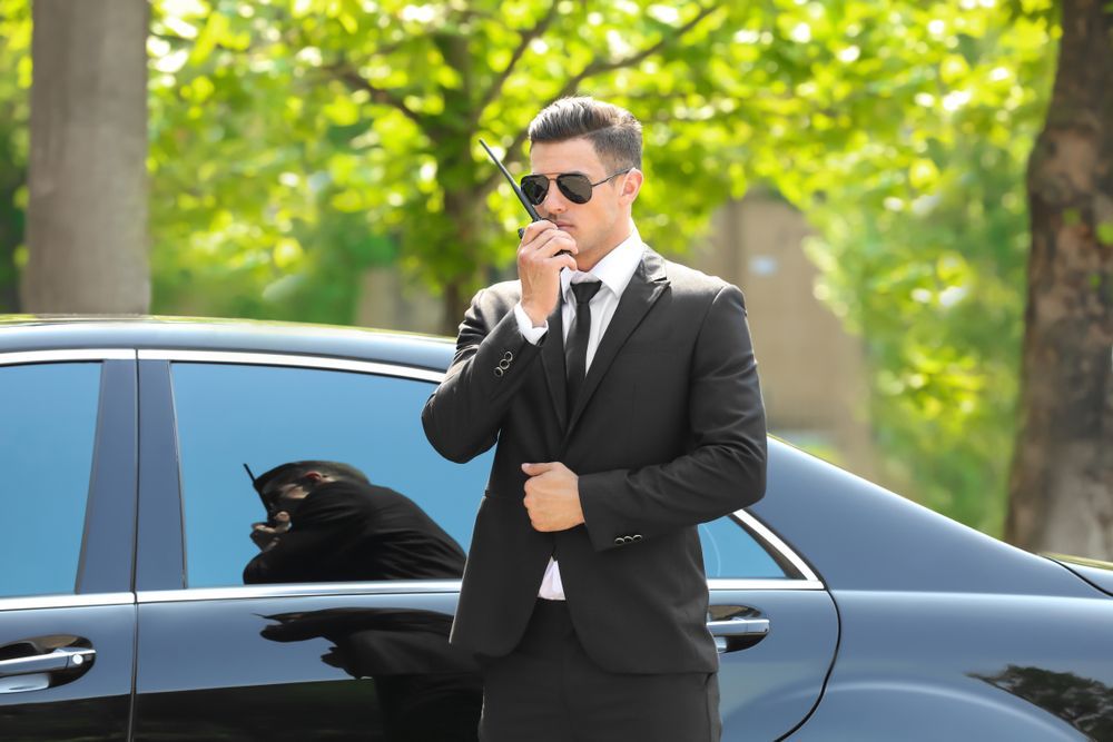 A man in a suit and tie is standing next to a car and talking on a walkie talkie.